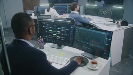 Male African American engineer types keyboard and analyzes displayed server data and real-time charts. Multi-cultural big data IT specialists work at background. Concept of cyber security. Back view.