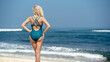 © asokova - A girl in a green swimsuit stands on the beach and looks at the ocean. View of a woman's back against the background of water and sand. Beach holidays in Bali. A blonde with tanned skin enjoys.
