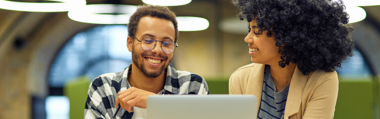 Two young happy multiracial business people sitting at the desk in the modern office, using laptop and discussing project, man and woman working together in coworking space