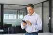 © insta_photos - Smiling older business man executive standing in office using mobile phone. Happy mid aged professional manager holding cell working on smartphone checking financial transactions on cellphone tech.