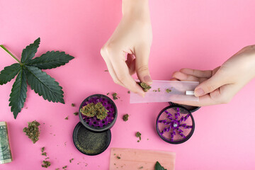 Wall Mural - Female hands twist a joint from dry buds of medical marijuana on a pink background.  Green leaves of cannabis lie nearby and the grinder is opened, pieces of weeds are around