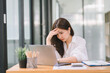 © kenchiro168 - The stressed and exhausted millennial businesswoman is seen sitting at her office desk with her hand on her head, indicating a hard working day where she is overloaded with work.