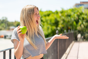 Wall Mural - Young blonde woman with an apple at outdoors with surprise facial expression