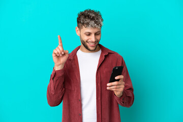 Wall Mural - Young handsome caucasian man isolated on blue background using mobile phone and lifting finger