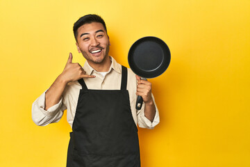 Asian chef holding a pan, yellow studio backdrop showing a mobile phone call gesture with fingers.