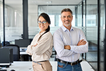 Wall Mural - Happy confident professional mature Latin business man and Asian business woman corporate leaders managers standing in office, two diverse colleagues executives team posing arms crossed, portrait.