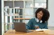 © crizzystudio - Young African businesswoman wearing headphones on her neck while looking at online shopping data Taking note of customer orders on a laptop while holding a mug of warm coffee to relax.
