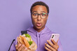 © WHstudio Leushin N - Surprised male student holds fast food sandwich and smartphone stands speechless stares bugged eyes wears transparent eyeglasses and hoodie carries rucksack isolated over purple studio background