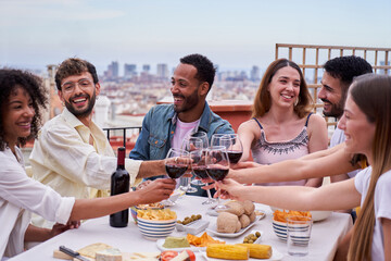 Multi-ethnic group of happy people toasting with red wine on a rooftop in the city. Friends having fun together, celebrating in a terrace during daytime, sitting at a table full of food. 