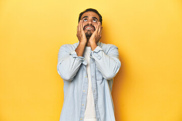 Wall Mural - Casual young Latino man against a vibrant yellow studio background, whining and crying disconsolately.