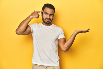 Wall Mural - Casual young Latino man against a vibrant yellow studio background, holding and showing a product on hand.