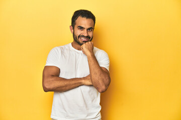 Wall Mural - Casual young Latino man against a vibrant yellow studio background, smiling happy and confident, touching chin with hand.