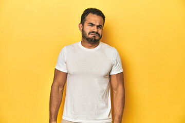 Wall Mural - Casual young Latino man against a vibrant yellow studio background, blows cheeks, has tired expression. Facial expression concept.