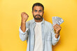Young Latino man holding a bundle of dollars, yellow studio background, showing fist to camera, aggressive facial expression.