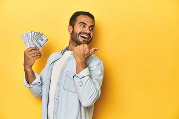 Young Latino man holding a bundle of dollars, yellow studio background, points with thumb finger away, laughing and carefree.