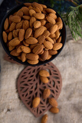 Wall Mural - Top view of a deep black bowl filled with dried healthy almonds against the background of old burlap and a green branch of thuja. Delicious raw almond nut in a bowl against a burlap cloth background