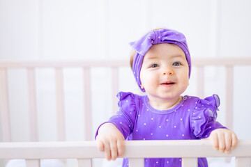 cute portrait of a baby girl in a crib holding on to the side in lilac clothes and with a bow on her head and laughing, the concept of children's goods