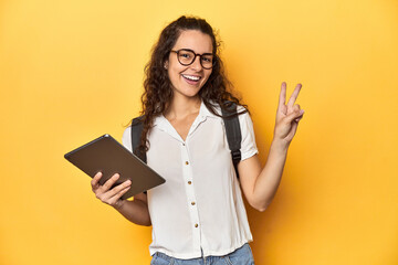 Student holding a tablet, glasses, backpack on, joyful and carefree showing a peace symbol with fingers.