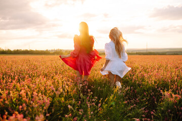 Two woman in stylish summer dresses feeling free in the field with flowers in sunshine. Nature, vacation, relax and lifestyle. Summer landscape.