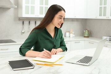 Canvas Print - Home workplace. Happy woman with pen and notebook working on laptop at marble desk in kitchen