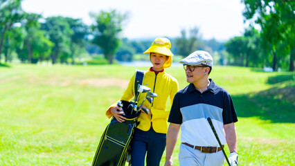 Asian man golfer walking together with female caddy on fairway at country club golf course. Healthy man enjoy outdoor activity lifestyle sport training golfing at golf club on summer holiday vacation.