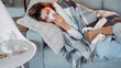 © golubovy - Home flu. Runny nose symptom. Suffering woman wrapped plaid laying sofa sneezing napkin feeling bad high temperature in light home interior.