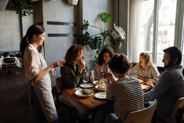 Wall Mural - Beautiful waitress serving group of cheerful friends in restaurant