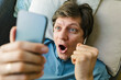 © Westend61 - Excited man holding smart phone lying down on bed at home
