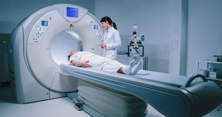 Female medical worker dressed up in gown before MRI examination. Female doctor is preparing patient for magnetic resonance procedure. Patient is lying at CT scanner bad and waiting to be scanned.