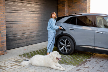 Young woman charges her electric car while standing with her cute dog near garage of her house. Concept of modern and happy lifestyle