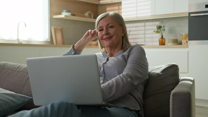 Relaxed happy elderly caucasian woman relaxing resting at home sofa typing on laptop smiling mature female browsing social network chatting online shopping in Internet watching movie use computer app