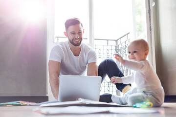 Wall Mural - Father working on laptop and watching baby daughter playing on floor