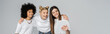 © LIGHTFIELD STUDIOS - Smiling interracial teenage girlfriends in white t-shirts using digital tablet together while hugging and standing isolated on grey, teenagers bonding over common interest, banner