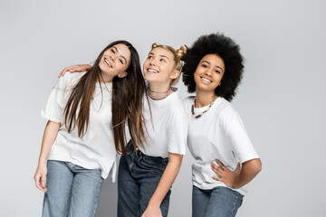 Multiethnic teenage girlfriends in jeans and casual white t-shirts posing while hugging and standing together isolated on grey, adolescence models and generation z concept, friendship