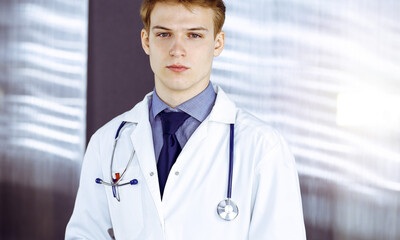 Wall Mural - Young doctor standing with crossed arms in sunny clinic near his working place. Medicine concept