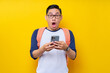 © Bangun Stock Photo - Shocked young Asian man student in casual clothes backpack glasses standing while holding mobile cell phone in hand and open mouth isolated on yellow background. high school university college concept
