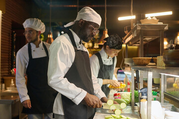 Young serious African American male cook in uniform chopping fresh zucchini for steaming while standing by counter among colleagues