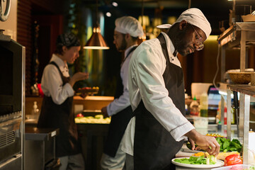 Young serious chef of restaurant mixing fresh chopped vegetables on round board while standing by workplace against two colleagues