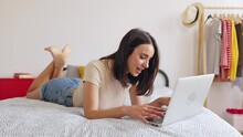Happy young woman using laptop lying on bed at home. Millennial teenage girl working or studying online on computer in bedroom. Technology and domestic life concept.