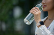 Young Asian woman drinking fresh water from a glass bottle outdoors. Thirsty female person taking a sip of mineral water on a walk
