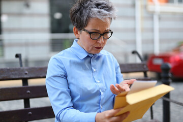 Poster - A stylish woman carefully reads a letter or notice from an envelope while sitting on a street bench.