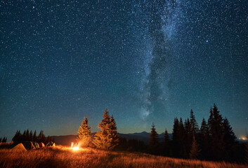 Night camping in mountains under starry sky. Tourist tents in campsite under sky full of stars with Milky way. Silhouette of hikers having a rest near burning campfire. Concept of tourism, adventures.