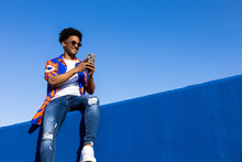 Smiling Black Man Using Smartphone And Sitting On Fence