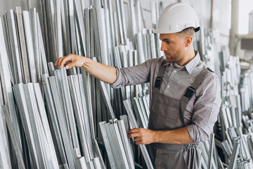 Happy bearded male factory worker in special uniform and white hard hat holding aluminum frame at production of metal plastic windows and doors