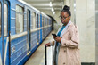 Young serious female business traveler with suitcase texting in smartphone while standing in front of camera with moving train on background