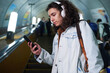 Teenage guy in casualwear and headphones using smartphone while standing on escalator, watching video and moving downwards