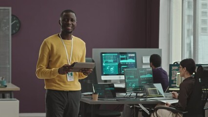 Medium portrait of young African American software engineer with digital tablet in hands smiling at camera standing in modern office with colleagues monitoring data on computers in background