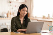 © Prostock-studio - Smiling young asian woman sitting at kitchen, using laptop