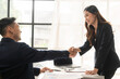 Happy young asian business man and woman shaking hands to congratulate successful collaboration on business deal with documents and laptop on table
