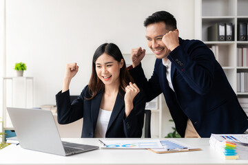 Two successful young Asian businessmen raising their hands in joy with laptop computers. ​​Celebrate victories and successes together. Clap hands in modern office